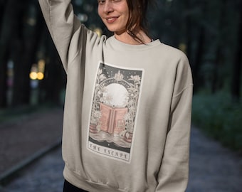 Puede incluir: Una sudadera beige con un estampado gráfico de una carta de tarot. La carta se titula "The Escape" y presenta un libro con flores y una luna en el fondo.