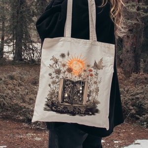 May include: A white canvas tote bag with a design of an open book with a sun and flowers growing out of it. The book is open to reveal a dark interior with glowing lights.