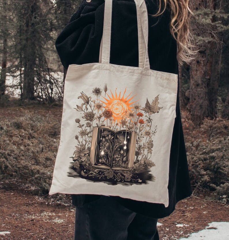 May include: A white canvas tote bag with a design of an open book with a sun and flowers growing out of it. The book is open to reveal a dark interior with glowing lights.