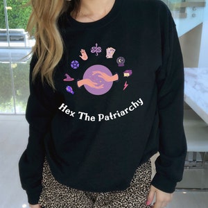 May include: Black sweatshirt featuring a graphic of hands holding a purple orb surrounded by various symbols. The text "Hex The Patriarchy" is printed below the graphic.