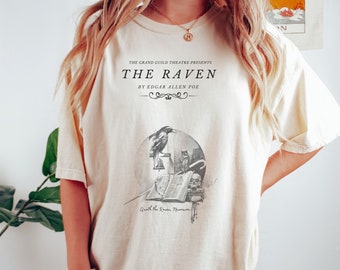 May include: A cream colored t-shirt with a black graphic print of a raven perched on a book. The text "THE RAVEN" is printed in black above the graphic. The text "BY EDGAR ALLEN POE" is printed in black below the graphic. The text "Quoth the Raven, Nevermore." is printed in black below the graphic.