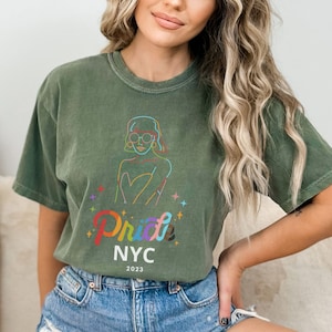 May include: A green t-shirt with a rainbow graphic of a person wearing glasses and the text "Pride NYC 2023".