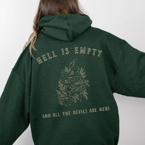 Puede incluir: Una sudadera con capucha verde oscuro con el texto "Hell is empty and all the devils are here" impreso en la espalda. El texto está en letras doradas y presenta una serpiente y flores.
