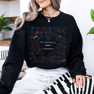 Pode incluir: Camisola de moletom preta com uma estampa gr&aacute;fica de selos de passaporte coloridos e o texto "Tudo o que voc&ecirc; precisa &eacute; amor - um passaporte".