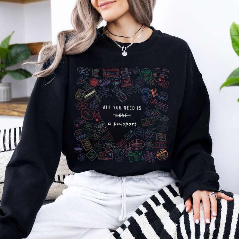 Pode incluir: Camisola de moletom preta com uma estampa gr&aacute;fica de selos de passaporte coloridos e o texto "Tudo o que voc&ecirc; precisa &eacute; amor - um passaporte".