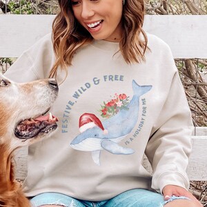 May include: Beige sweatshirt with a watercolor whale design. The whale is light blue and wears a Santa hat, with a bouquet of flowers on its back. The text reads "FESTIVE, WILD & FREE IS A HOLIDAY FOR ME."