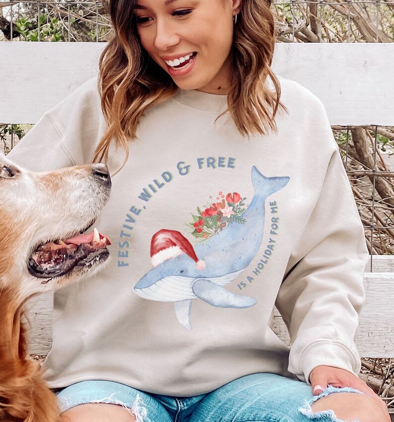May include: Beige sweatshirt with a watercolor whale design. The whale is light blue and wears a Santa hat, with a bouquet of flowers on its back. The text reads "FESTIVE, WILD & FREE IS A HOLIDAY FOR ME."