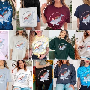 May include: Collection of apparel featuring a whimsical whale design. The whale wears a Santa hat and carries flowers, with the text "FESTIVE, WILD & FREE" and "IS A HOLIDAY FOR ME". Includes t-shirts, sweatshirts, and tote bags in various colors and tie-dye.