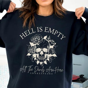 Puede incluir: Una sudadera negra con un diseño de calavera blanca y flores. El texto "Hell is Empty All The Devils Are Here Shakespeare" está impreso en la parte delantera.