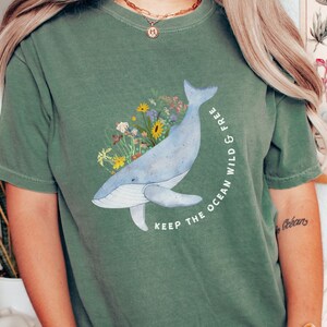 以下が含まれることがあります： 青いクジラとカラフルな花が描かれた緑色のTシャツ。Tシャツには「Keep the ocean wild & free」という文字がプリントされています。
