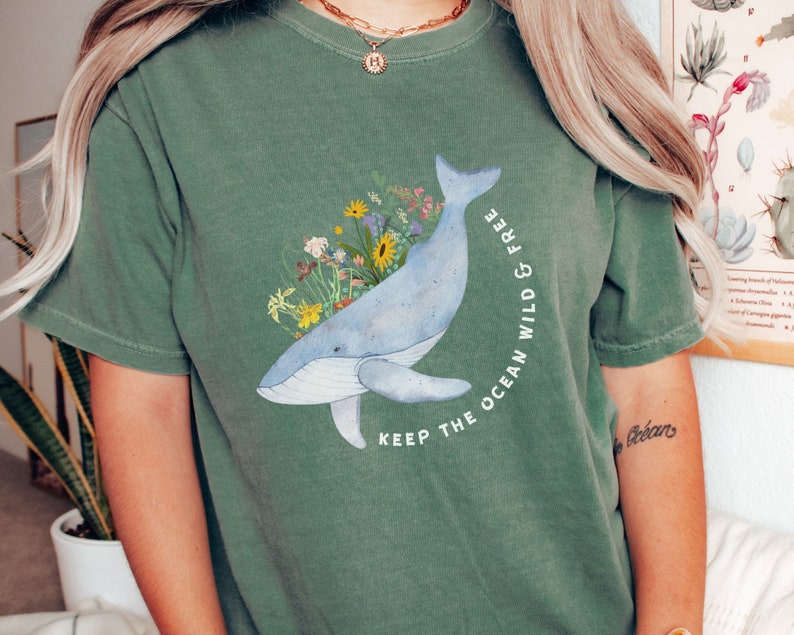 以下が含まれることがあります： 青いクジラとカラフルな花が描かれた緑色のTシャツ。Tシャツには「Keep the ocean wild & free」という文字がプリントされています。