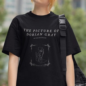 May include: Black t-shirt with a white graphic design featuring a line drawing of a face with the text "THE PICTURE OF DORIAN GRAY BY OSCAR WILDE" and "TO DEFIKE IS TO LIMIT".