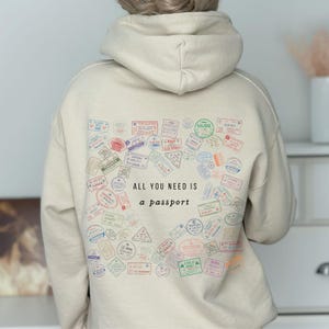 May include: Beige hoodie with a passport stamp design. The back of the hoodie features a collection of colorful passport stamps and the text "ALL YOU NEED IS a passport". The stamps are in various shapes and sizes.