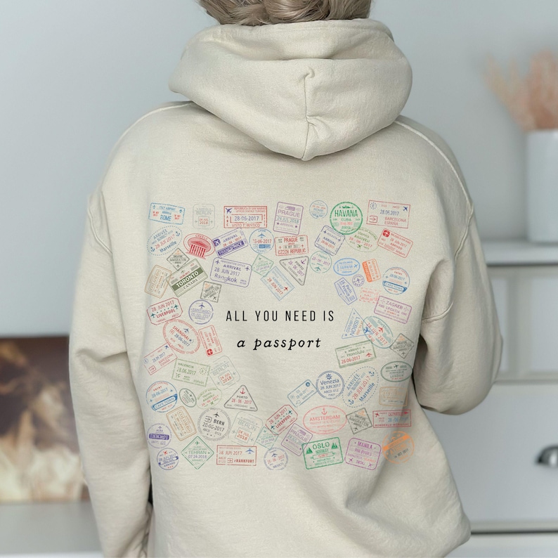 May include: Beige hoodie with a passport stamp design. The back of the hoodie features a collection of colorful passport stamps and the text "ALL YOU NEED IS a passport". The stamps are in various shapes and sizes.