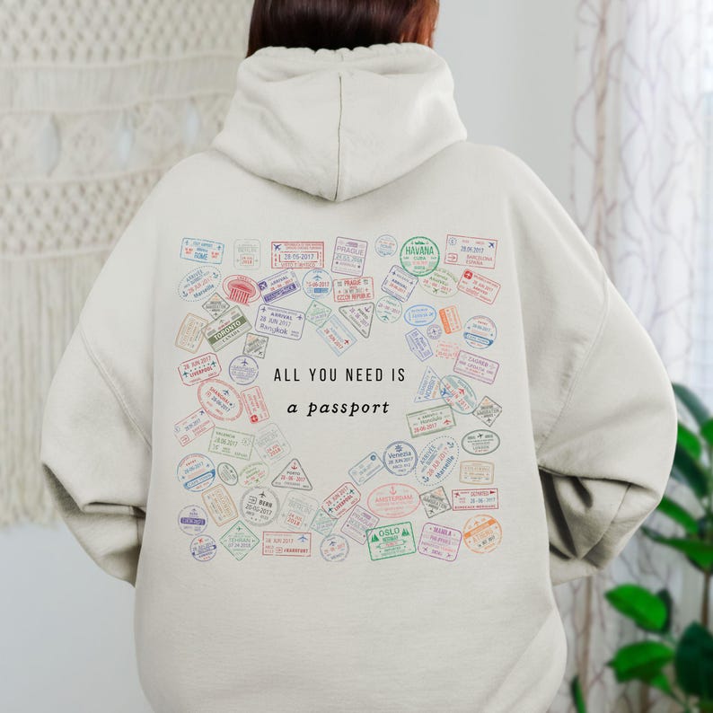 May include: Cream-colored hoodie featuring a collection of colorful passport stamps. The phrase "ALL YOU NEED IS a passport" is printed in black. The hoodie has a hood and long sleeves.