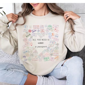 Pode incluir: Uma camisa de moletom cor creme com uma estampa gr&aacute;fica de carimbos de passaporte e o texto "All you need is love + a passport".