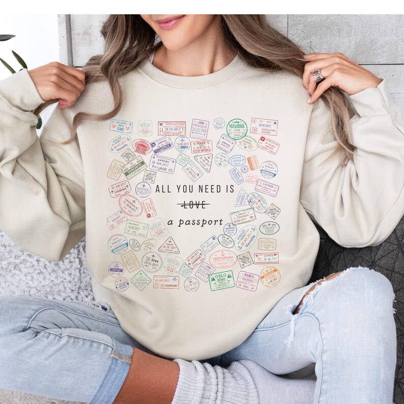 Pode incluir: Uma camisa de moletom cor creme com uma estampa gr&aacute;fica de carimbos de passaporte e o texto "All you need is love + a passport".