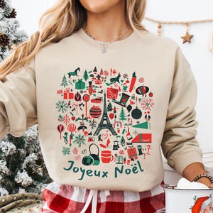以下が含まれることがあります： クリスマスのモチーフがデザインされたベージュのトレーナー。赤い星、緑のクリスマスツリー、プレゼント、トナカイなど、赤と緑のアイコンが多数描かれています。デザインの下には「Joyeux Noël」という文字が緑色で印刷されています。