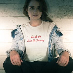 May include: A person wearing a white t-shirt with a red cherry graphic and the text "Smash The Patriarchy". They are wearing a denim jacket and black pants.