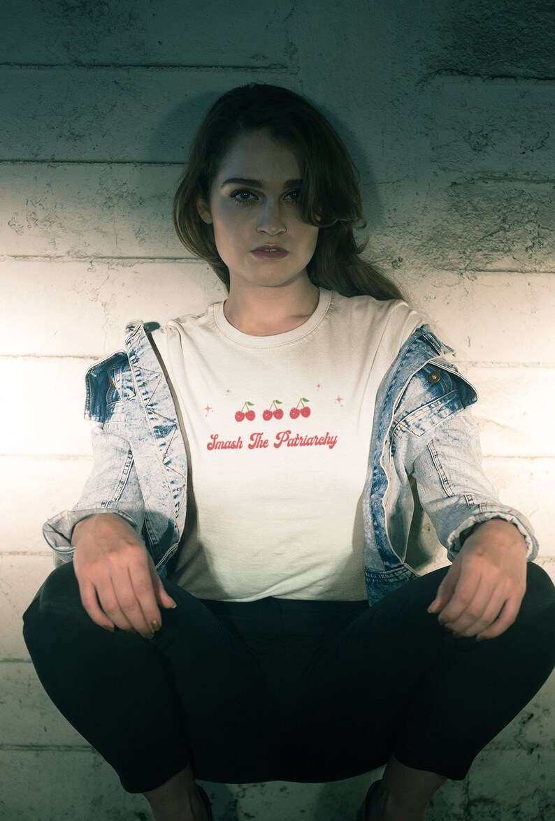 May include: A person wearing a white t-shirt with a red cherry graphic and the text "Smash The Patriarchy". They are wearing a denim jacket and black pants.