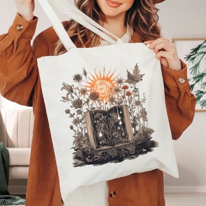 May include: A white canvas tote bag with a colorful illustration of a book surrounded by flowers and a sun. The book is open and has a glowing light emanating from it.