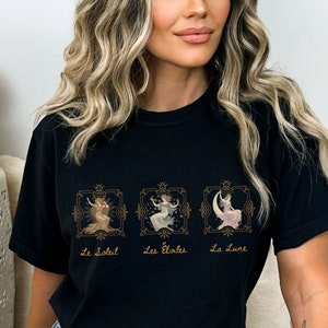 May include: Black t-shirt with a graphic of three women in gold frames. The text below the frames reads "Le Soleil", "Les Etoiles", and "La Lune".