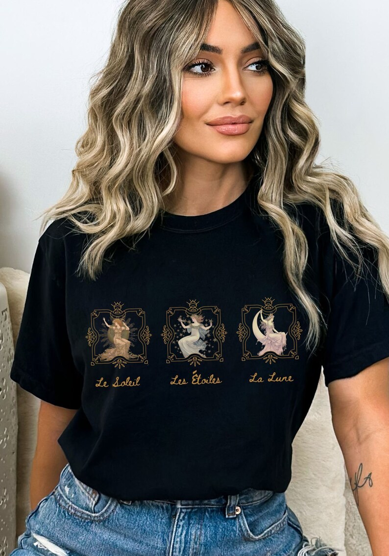 May include: Black t-shirt with a graphic of three women in gold frames. The text below the frames reads "Le Soleil", "Les Etoiles", and "La Lune".