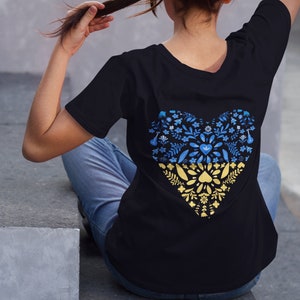 May include: Black t-shirt featuring a heart-shaped design on the back, filled with blue and yellow floral patterns. The design incorporates elements of leaves and flowers, creating a decorative and symbolic image. The shirt is a casual wear item.