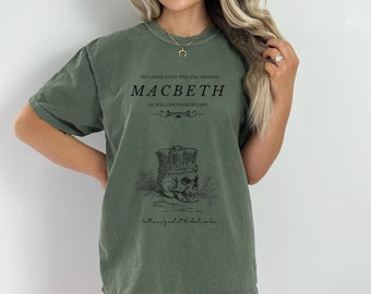 May include: A green t-shirt with a graphic print of a skull wearing a crown. The text on the shirt reads "THE GRAND GUILD THEATRE PRESENTS MACBETH BY WILLIAM SHAKESPEARE Hell is empty and all the devils are here."