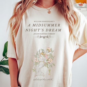 May include: Cream-colored t-shirt featuring the text "WILLIAM SHAKESPEARE'S A MIDSUMMER NIGHT'S DREAM AN ENCHANTED COMEDY." Below the text is a floral illustration with a figure. The quote "Lord what fools these mortals be" is at the bottom.