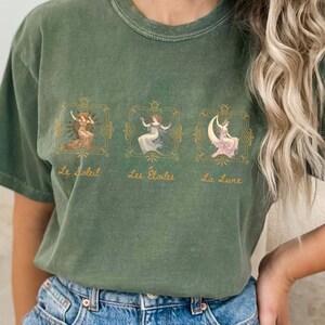 May include: A green t-shirt with a graphic print of three women in gold frames. The text "Le Soleil", "Les Etoiles", and "La Lune" is printed below the frames.