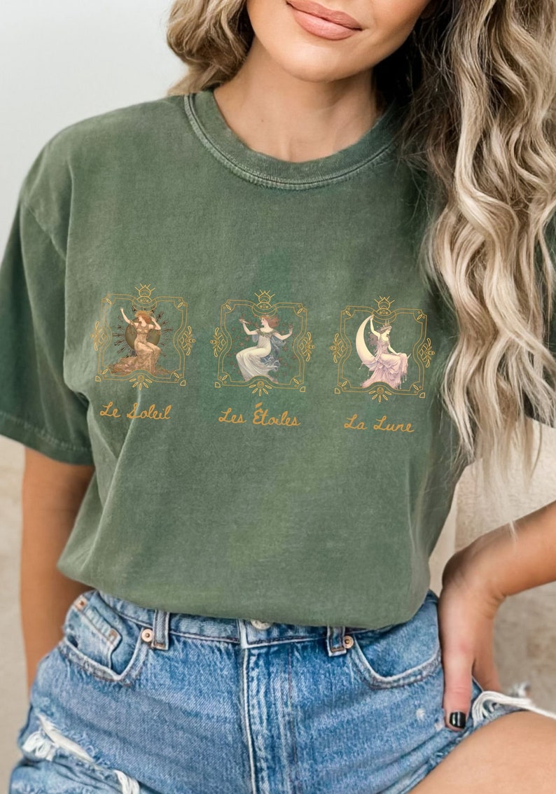 May include: A green t-shirt with a graphic print of three women in gold frames. The text "Le Soleil", "Les Etoiles", and "La Lune" is printed below the frames.