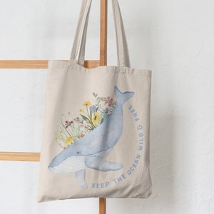 May include: A beige tote bag featuring a blue whale illustration with floral accents. The whale is surrounded by colourful flowers and the text "Keep the ocean wild & free" is printed on the bag.