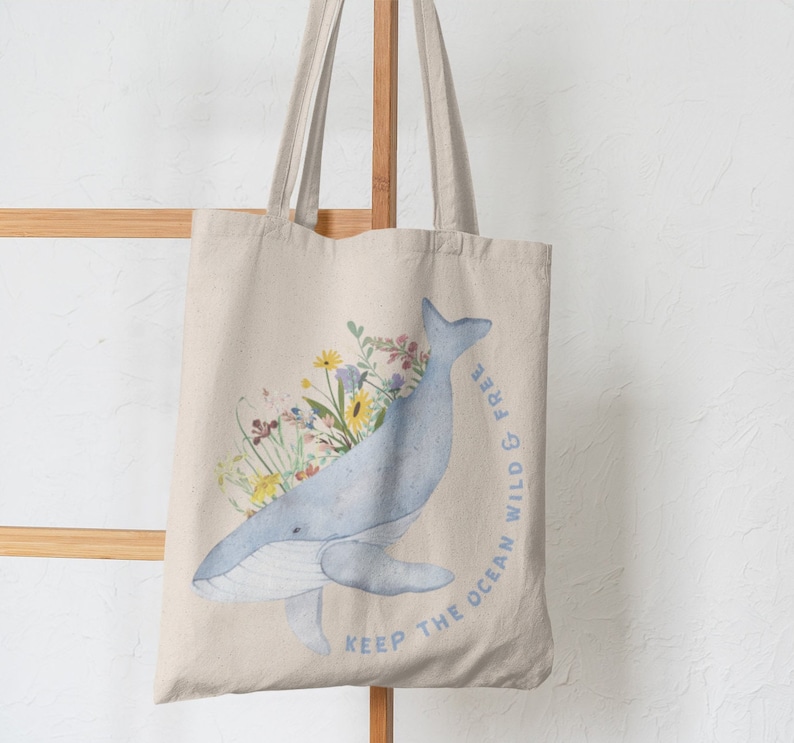 May include: A beige tote bag featuring a blue whale illustration with floral accents. The whale is surrounded by colourful flowers and the text "Keep the ocean wild & free" is printed on the bag.