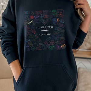 May include: Black hooded sweatshirt with a graphic print of colorful passport stamps and the text "All you need is love + a passport".