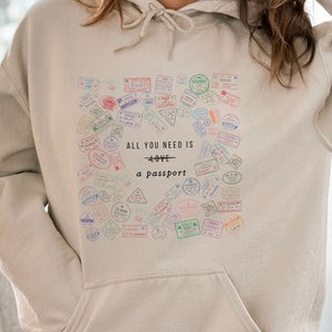 May include: A beige hoodie with a heart-shaped design featuring a collection of colorful passport stamps. The text "ALL YOU NEED IS LOVE - a passport" is printed on the hoodie.