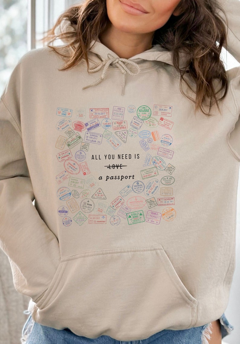 May include: A beige hoodie with a heart-shaped design featuring a collection of colorful passport stamps. The text "ALL YOU NEED IS LOVE - a passport" is printed on the hoodie.