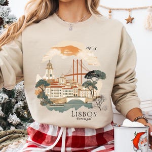 May include: A beige sweatshirt with a graphic print of the Lisbon skyline, including the 25 de Abril Bridge, a church, and buildings. The text "LISBON Portugal" is printed below the graphic.