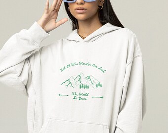 May include: A light gray hoodie with a front pocket and a drawstring hood. The hoodie features a green graphic of mountains and trees with the text "Not All Who Wander Are Lost" and "The World Is Yours". The person is wearing blue sunglasses.