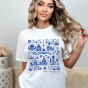 K&ouml;nnte beinhalten: Wei&szlig;es T-Shirt mit einem blauen und wei&szlig;en Delft, Zuid-Holland, Niederlande Design. Das Design zeigt eine Vielzahl von niederl&auml;ndisch inspirierten Bildern, darunter H&auml;user, Windm&uuml;hlen, Blumen und Boote.