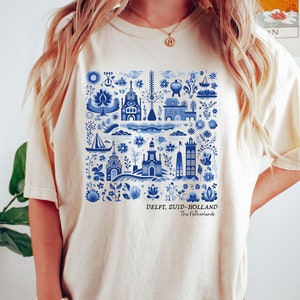 K&ouml;nnte beinhalten: Ein cremefarbenes T-Shirt mit einem blauen und wei&szlig;en Delft, Zuid-Holland, Niederlande Design. Das Design zeigt eine Vielzahl traditioneller niederl&auml;ndischer Bilder, darunter H&auml;user, Windm&uuml;hlen und Blumen.
