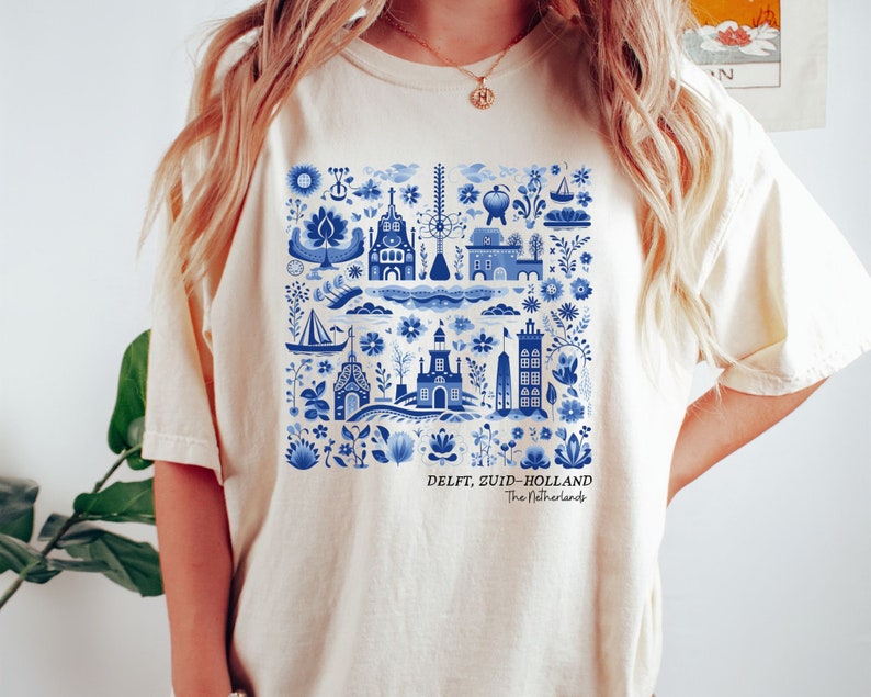 K&ouml;nnte beinhalten: Ein cremefarbenes T-Shirt mit einem blauen und wei&szlig;en Delft, Zuid-Holland, Niederlande Design. Das Design zeigt eine Vielzahl traditioneller niederl&auml;ndischer Bilder, darunter H&auml;user, Windm&uuml;hlen und Blumen.