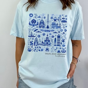 K&ouml;nnte beinhalten: Hellblaues T-Shirt mit einem blauen und wei&szlig;en Delft, Zuid-Holland, Niederlande Design. Das Design zeigt eine Vielzahl von Geb&auml;uden, Blumen und anderen Elementen.