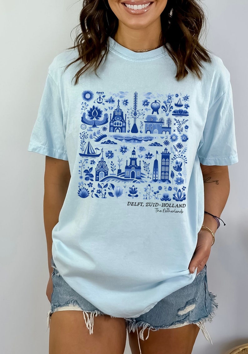 K&ouml;nnte beinhalten: Hellblaues T-Shirt mit einem blauen und wei&szlig;en Delft, Zuid-Holland, Niederlande Design. Das Design zeigt eine Vielzahl von Geb&auml;uden, Blumen und anderen Elementen.