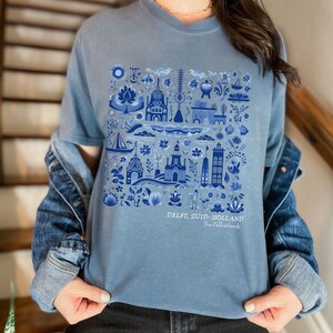 K&ouml;nnte beinhalten: Ein hellblaues T-Shirt mit einem blauen und wei&szlig;en Delft, Zuid-Holland, Niederlande Design. Das Design zeigt eine Vielzahl traditioneller niederl&auml;ndischer Bilder, darunter Windm&uuml;hlen, H&auml;user und Blumen.