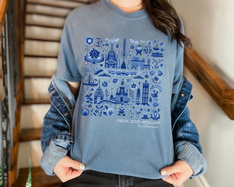 K&ouml;nnte beinhalten: Ein hellblaues T-Shirt mit einem blauen und wei&szlig;en Delft, Zuid-Holland, Niederlande Design. Das Design zeigt eine Vielzahl traditioneller niederl&auml;ndischer Bilder, darunter Windm&uuml;hlen, H&auml;user und Blumen.