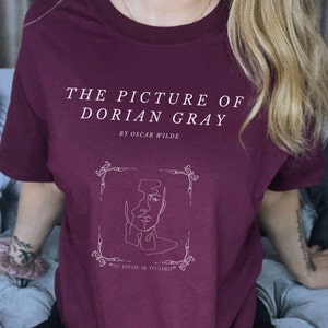 May include: A maroon t-shirt with a white graphic design featuring the title "The Picture of Dorian Gray" by Oscar Wilde. The graphic also includes a line drawing of a face and the quote "To define is to limit."