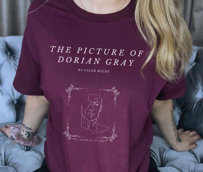 May include: A maroon t-shirt with a white graphic design featuring the title "The Picture of Dorian Gray" by Oscar Wilde. The graphic also includes a line drawing of a face and the quote "To define is to limit."