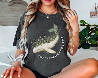 Puede incluir: Una camiseta gris oscuro con un gráfico de ballena blanca y detalles florales. El texto "Keep the ocean wild & free" está impreso en la camiseta.