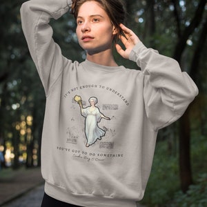 May include: A gray sweatshirt with a quote from author Flannery O'Connor. The quote reads "It's not enough to understand. You've got to do something." The quote is printed in black text on a white background. The sweatshirt features a graphic of a woman holding a book and a quill pen.